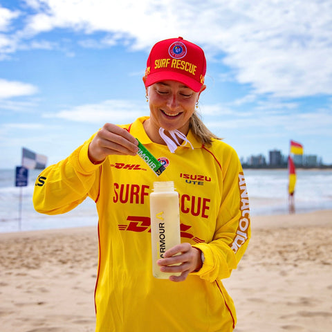 Person in yellow Surf Rescue uniform on a beach holding a bottle and snack.
