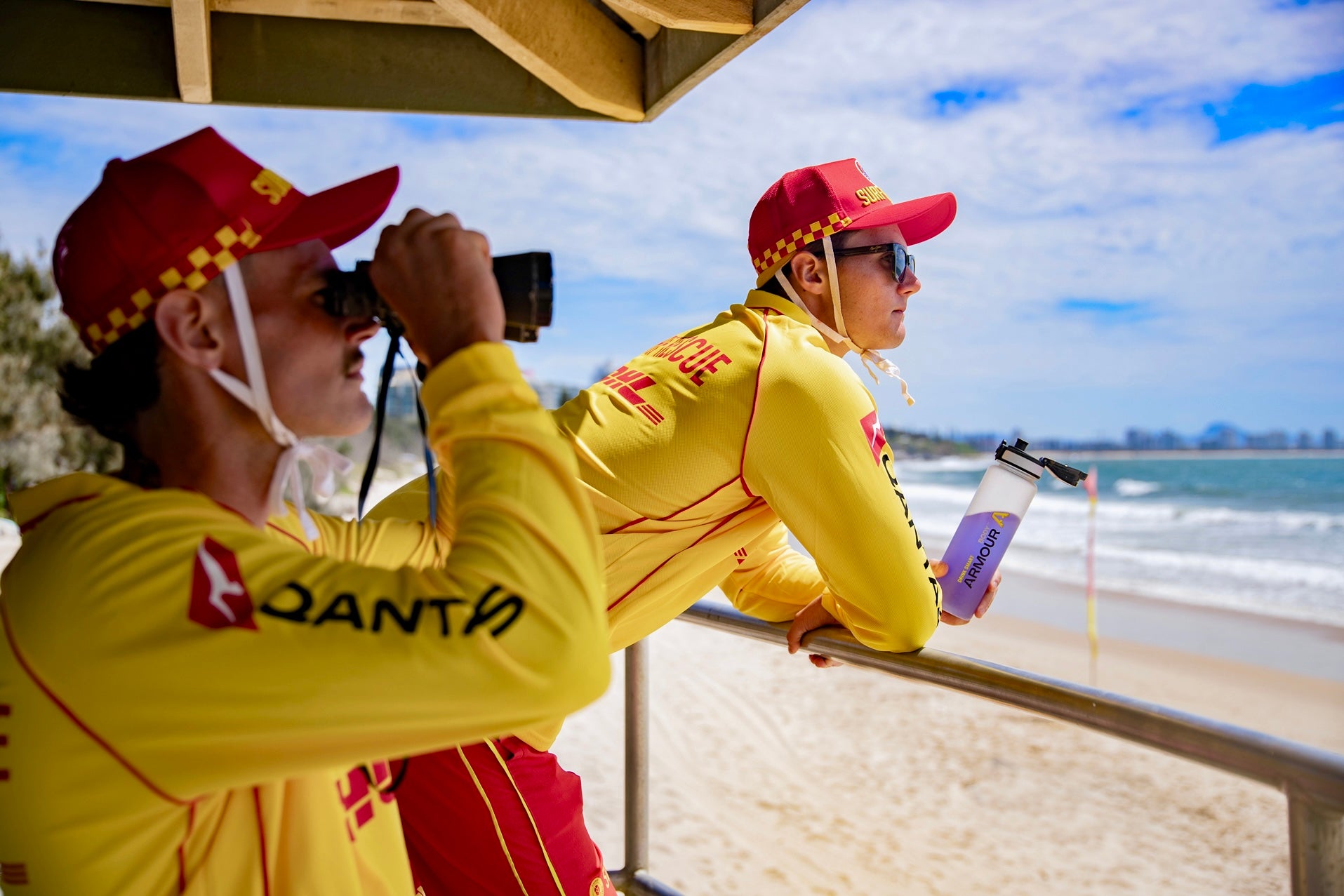 Two lifeguards in yellow uniforms with red hats on a beach, one using binoculars.